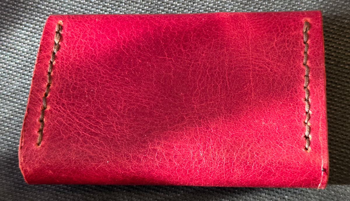 A close-up of a bright red leather wallet or card holder with visible grain texture and decorative contrast stitching on two sides, placed on a dark textured fabric background.