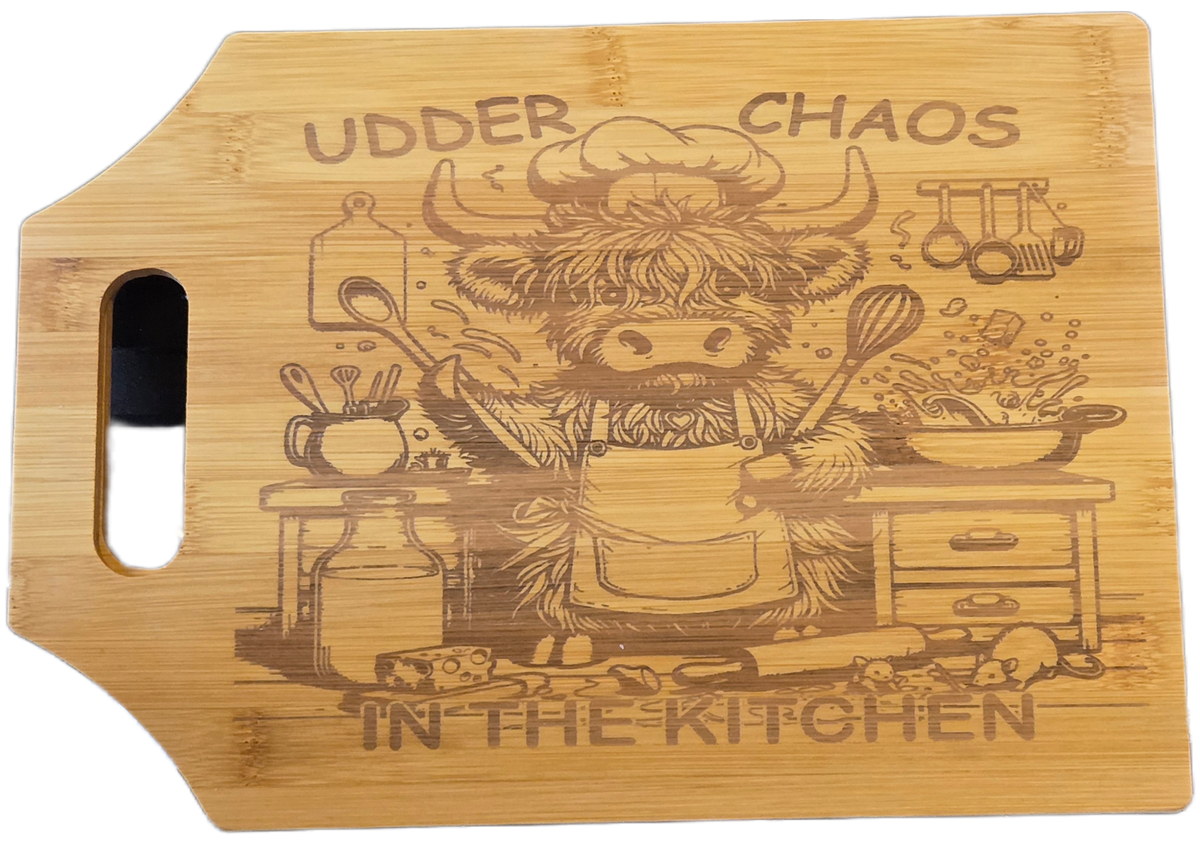 A rectangular bamboo cutting board with a handle cutout on the left, featuring a laser-engraved illustration of a funny Scottish highland cow in a chef's hat and apron cooking in a kitchen, with the text 'UDDER CHAOS' above and 'IN THE KITCHEN' below.
