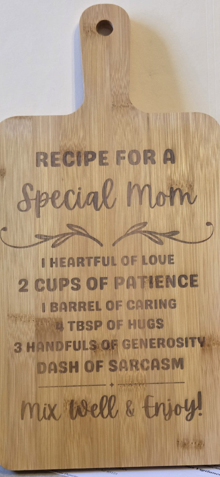 A rectangular wooden cutting board with a handle, featuring engraved text that reads 'RECIPE FOR A Special Mom' followed by sentimental phrases, photographed on a light countertop.