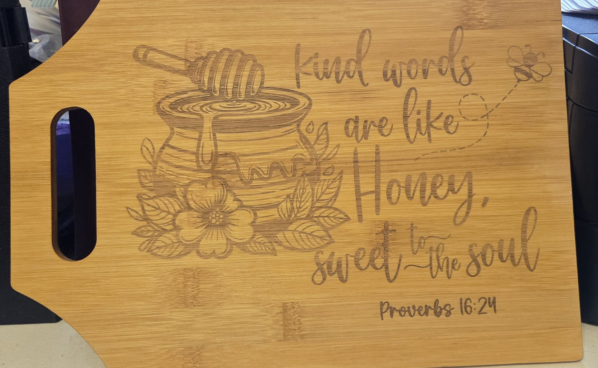 A rectangular wooden cutting board with a handle, featuring a laser-engraved design of a honey pot, flower, and bee with the quote 'Kind words are like Honey, sweet to the soul' and 'Proverbs 16:24'.