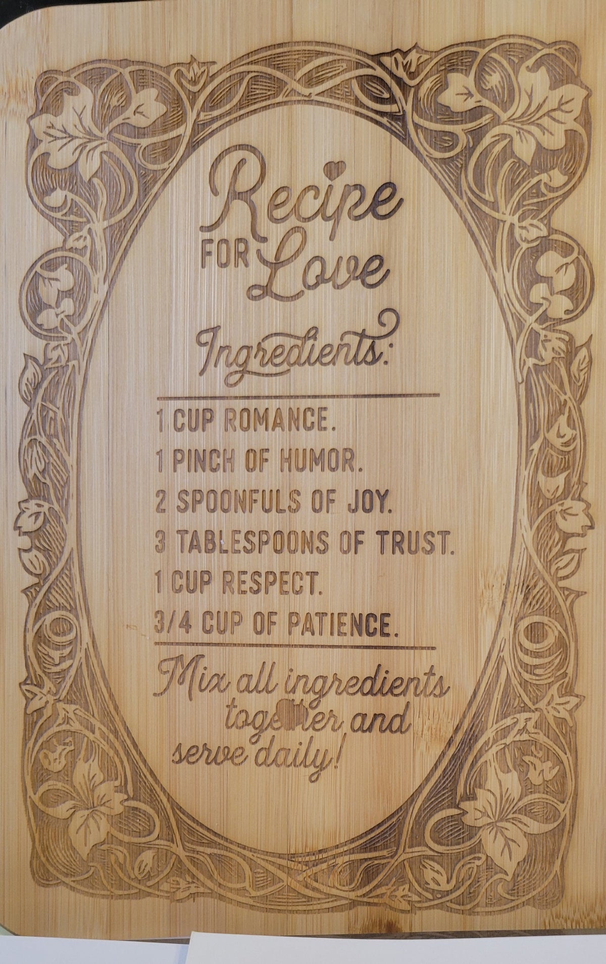 A rectangular wooden plaque or sign with engraved decorative floral borders and the text 'Recipe for Love Ingredients' followed by a humorous recipe with phrases like '1 cup romance' and 'mix all ingredients together and serve daily!'. The wood has a light natural tone with darker brown engraved lines.