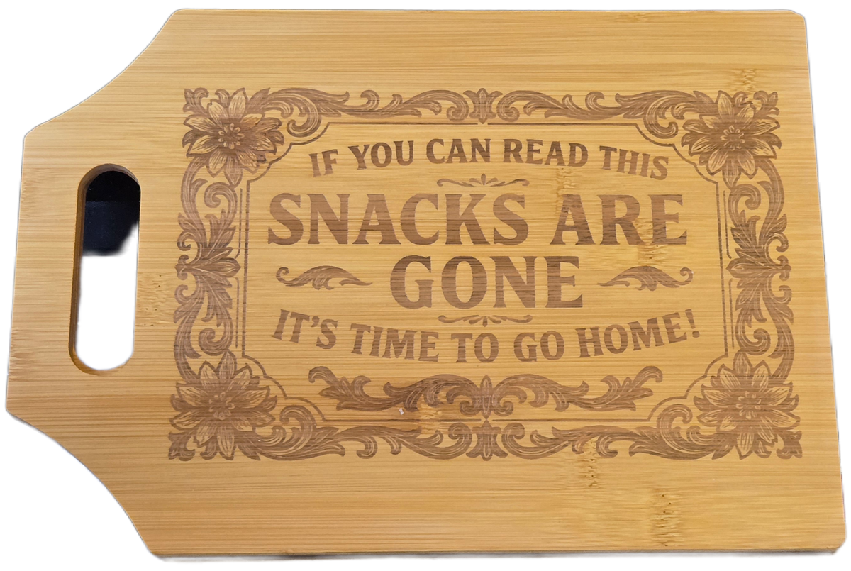 A rectangular bamboo cutting board with a die-cut handle on the left, featuring a laser-engraved decorative border and the humorous phrase 'IF YOU CAN READ THIS SNACKS ARE GONE IT'S TIME TO GO HOME!'.